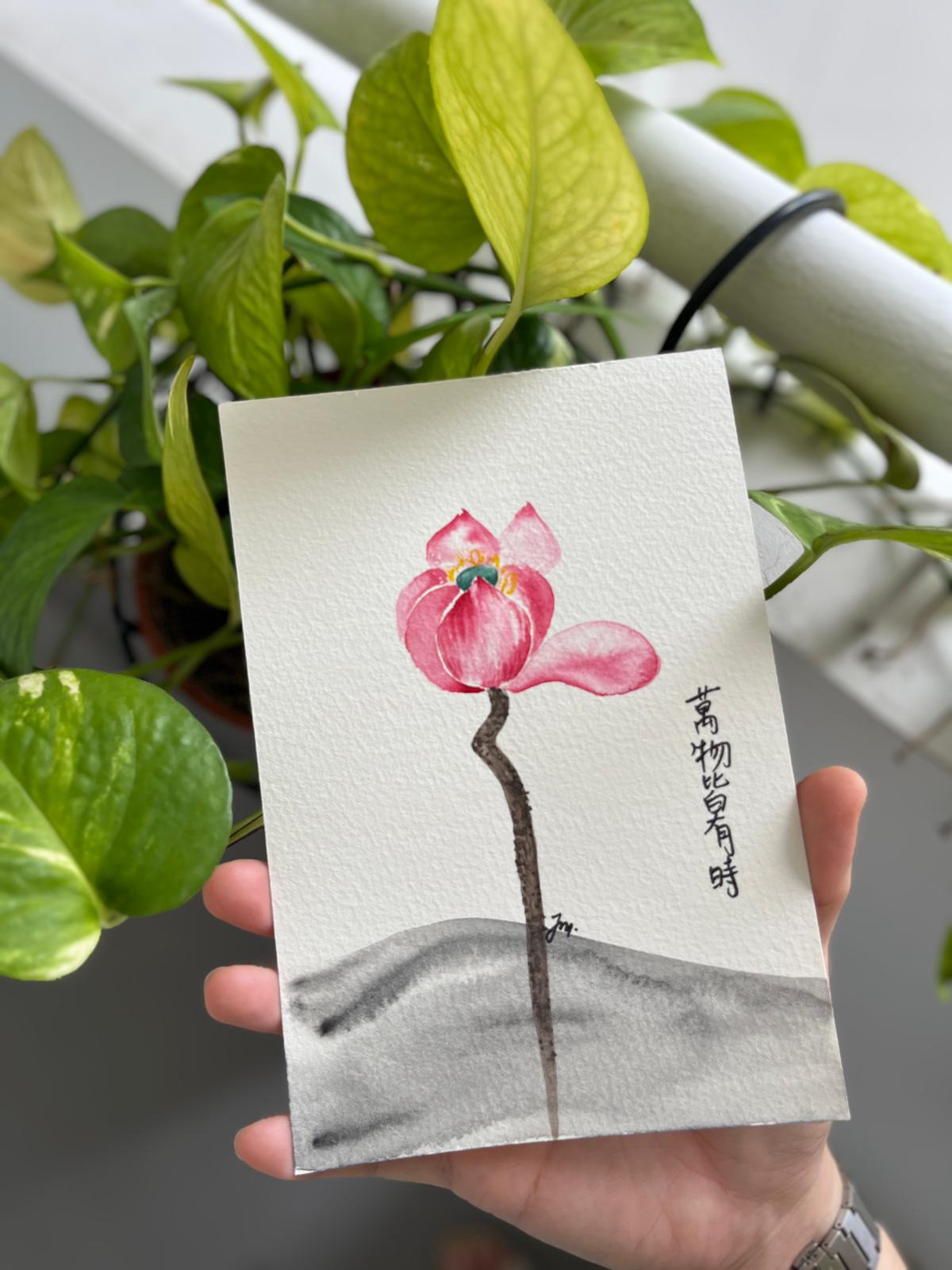 Watercolour painting of a pink Japanese Anemone with Chinese calligraphy, held up against a green plant backdrop